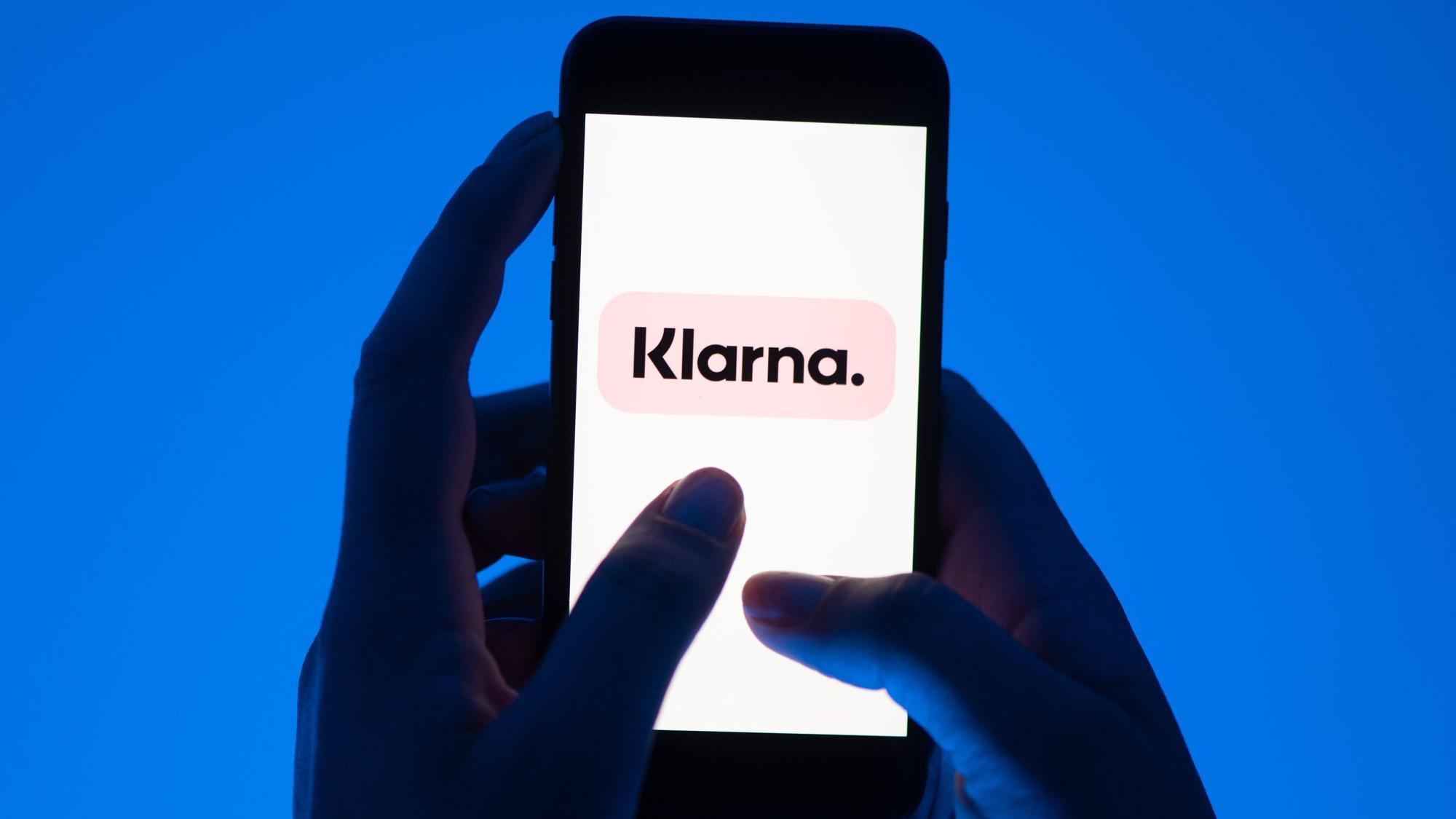 Zwei Hände halten ein Smartphone vor blauem Hintergrund, darauf ist groß das Klarna-Logo zu sehen.