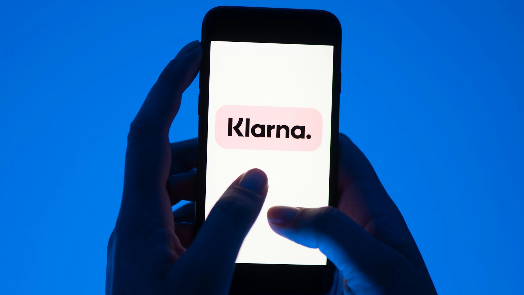 Zwei Hände halten ein Smartphone vor blauem Hintergrund, darauf ist groß das Klarna-Logo zu sehen.