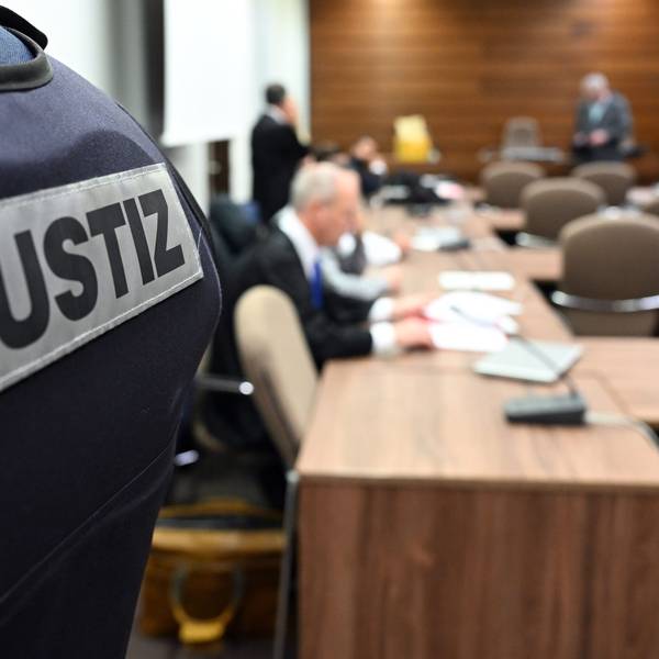 Im Vordergrund ist der Rücken eines Beamten mit der Aufschrift „Justiz“ zu sehen, im Hintergrund ist unscharf der Sitzungssaal im Kölner Landgericht.