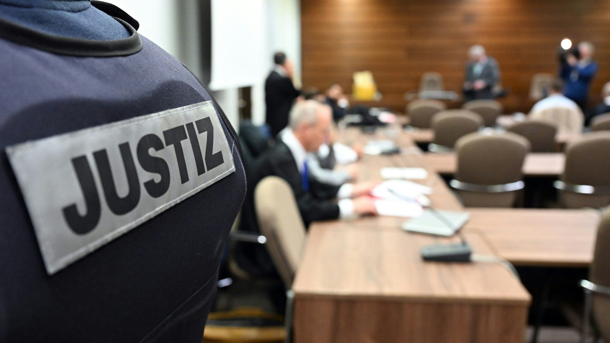 Im Vordergrund ist der Rücken eines Beamten mit der Aufschrift „Justiz“ zu sehen, im Hintergrund ist unscharf der Sitzungssaal im Kölner Landgericht.