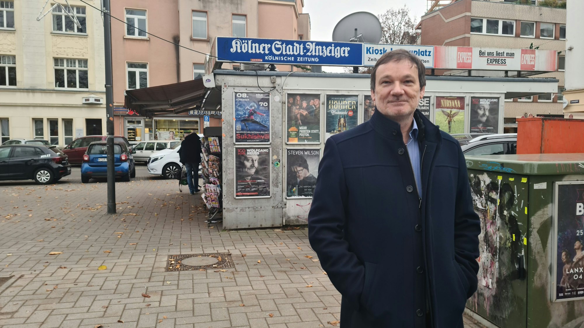 Das Bild zeigt einen Mann vor einem Kiosk auf einem Platz.