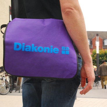 Ein Mann trägt eine violette Umhängetasche mit der Aufschrift Diakonie. im Hintergrund sind Menschen in der Troisdorfer Fußgängerzone zu sehen.