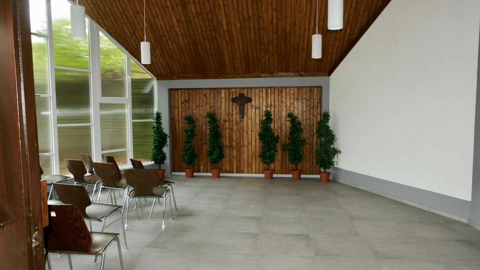 Ein Raum mit grauem Betonboden und einer getäfelten Decke ist zu sehen, lins ist eine Fensterfront, ein paar Stühle stehen an der Seite.