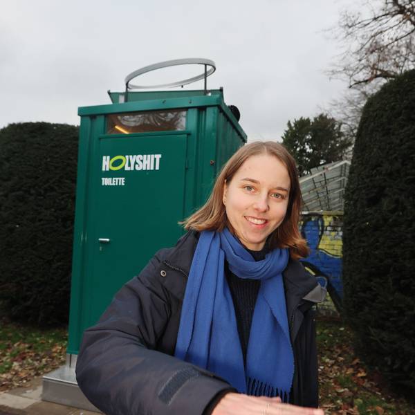 Anastasia Bondar steht vor einem grünen Toilettenhäuschen mit der Aufschrift „Holy Shit“.