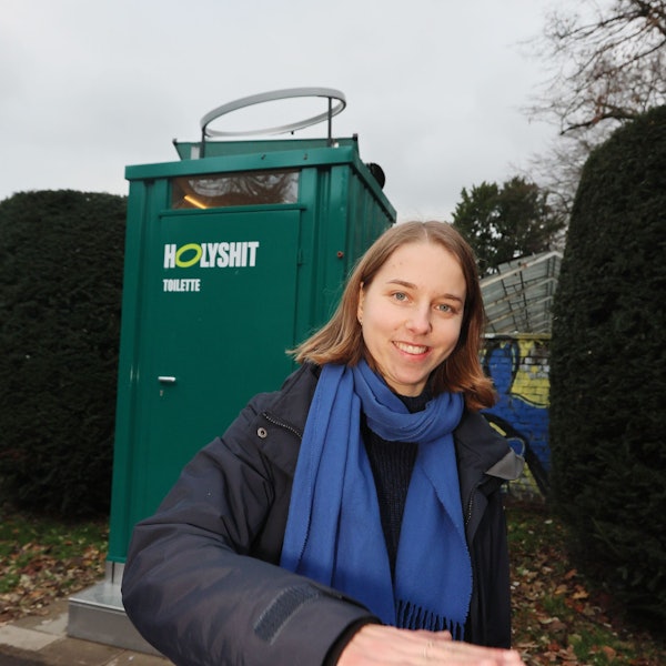 Anastasia Bondar steht vor einem grünen Toilettenhäuschen mit der Aufschrift „Holy Shit“.