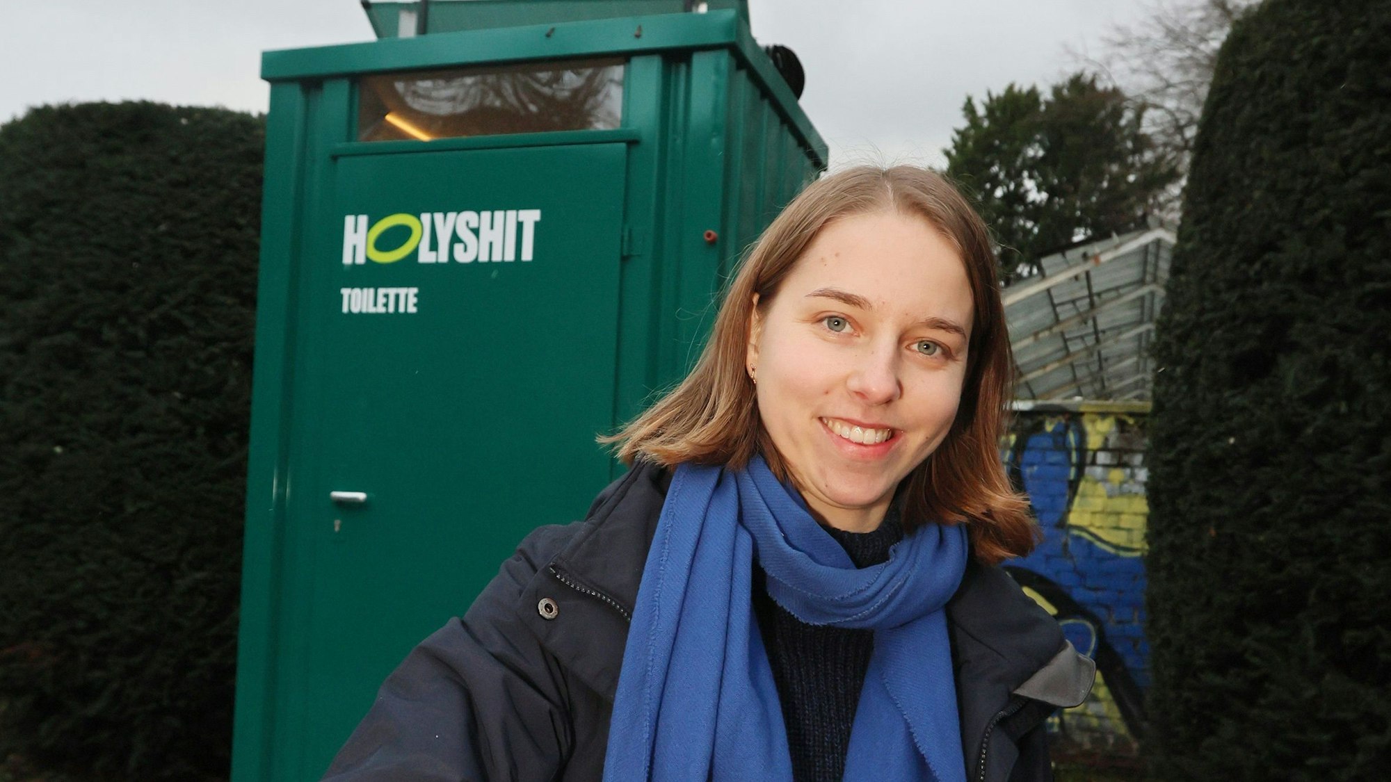 Anastasia Bondar steht vor einem grünen Toilettenhäuschen mit der Aufschrift „Holy Shit“.