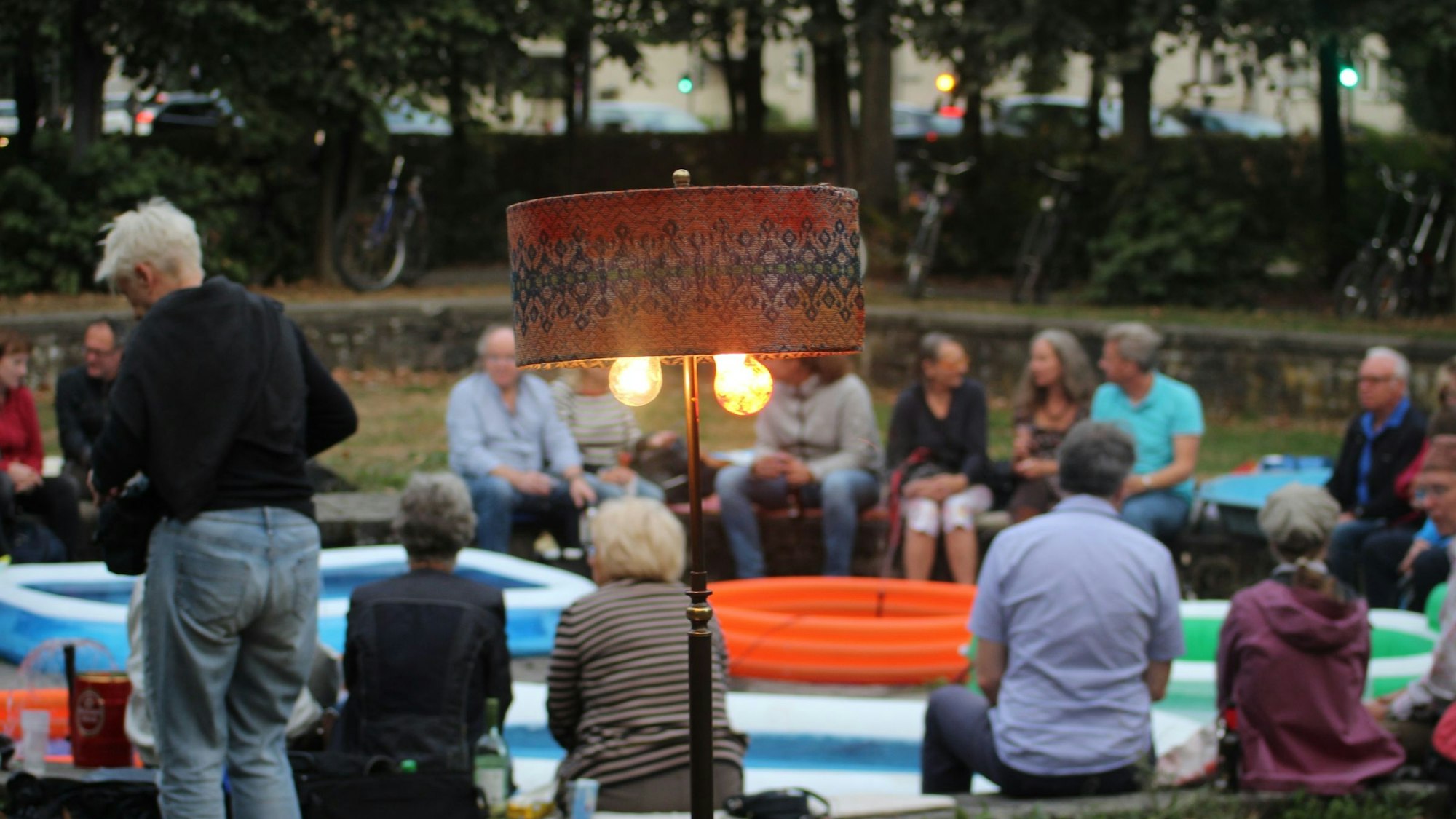 Menschen sitzen auf einem kreisförmigen Mäuerchen, im Kreis stehen mehrere Planschbecken.