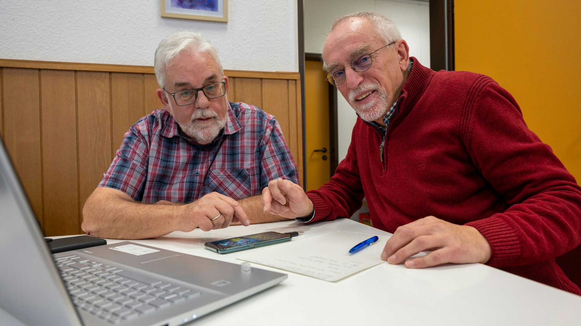 Das Smartcafé ist eine Seniorenbegegnungsstätte. Senioren fühlen sich von der Digitalisierung abgeschnitten. Zwei betagte Männer sitzen über ein Smartphone gebeugt gemeinsam am Tisch. Vor ihnen steht zudem ein aufgeklapper Laptop.