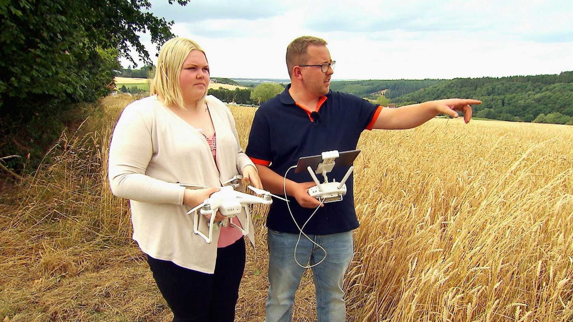 Stephan und Laura stehen mit einer Drohne vor einem sommerlichen Getreidefeld, Stephan deutet in die Ferne.