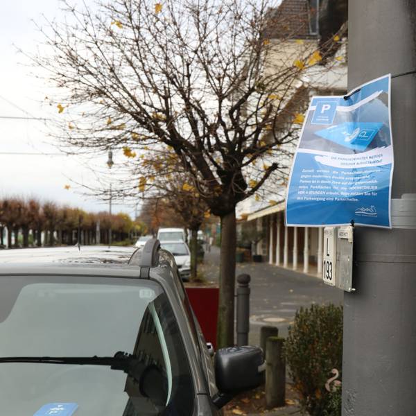 An einem Laternenpfahl neben parkenden Autos hängt ein laminiertes blaues Schild, auf dem die Stadt Autofahrer bittet, die Parkscheibe hinter die Windschutzscheibe zu legen.