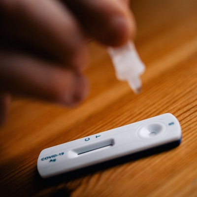 Ein Corona-Schnelltest liegt auf einem Holztisch. Eine Hand hält die kleine Testampulle.