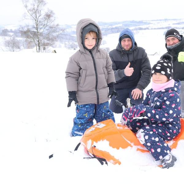 Zwei Kinder und zwei Männer beim Rodeln im Schnee. Die Kleinste sitzt auf einem orange farbigen Schlitten.