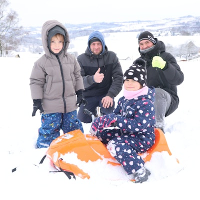 Zwei Kinder und zwei Männer beim Rodeln im Schnee. Die Kleinste sitzt auf einem orange farbigen Schlitten.