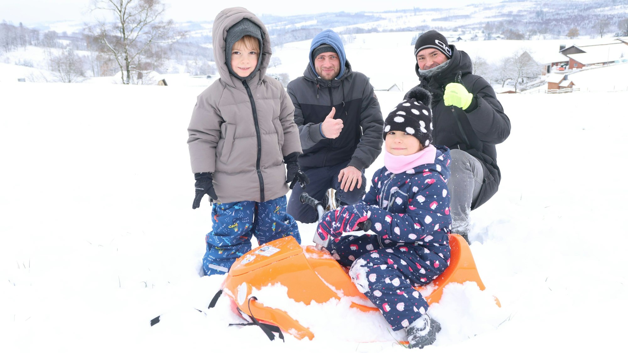 Zwei Kinder und zwei Männer beim Rodeln im Schnee. Die Kleinste sitzt auf einem orange farbigen Schlitten.