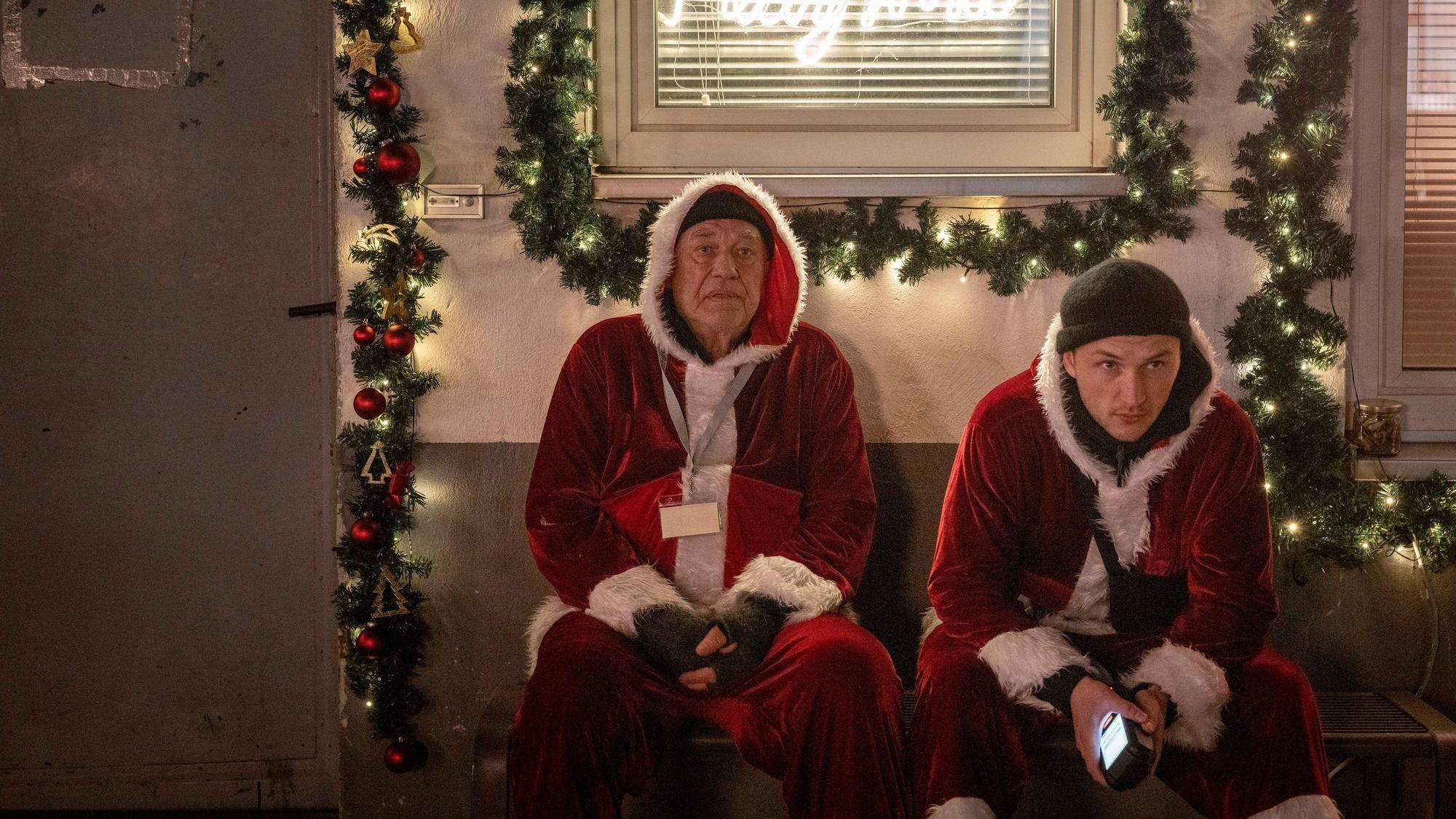 Zu sehen sind zwei Männer mit Weihnachtsmannkostümen. Sie sitzen nebeneinander. Hinter ihnen ist ihre Firma, sie ist weihnachtlich dekoriert.