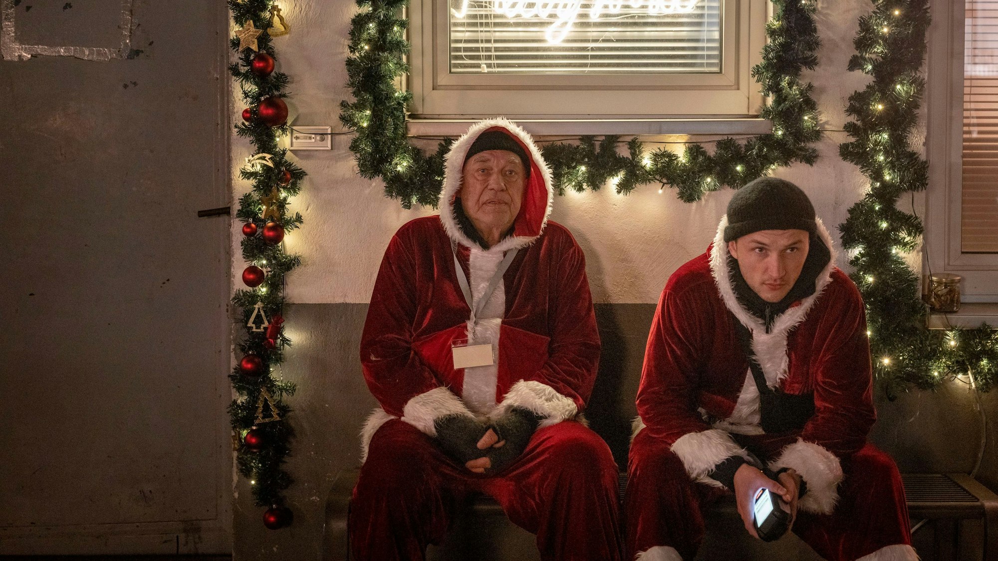 Zu sehen sind zwei Männer mit Weihnachtsmannkostümen. Sie sitzen nebeneinander. Hinter ihnen ist ihre Firma, sie ist weihnachtlich dekoriert.