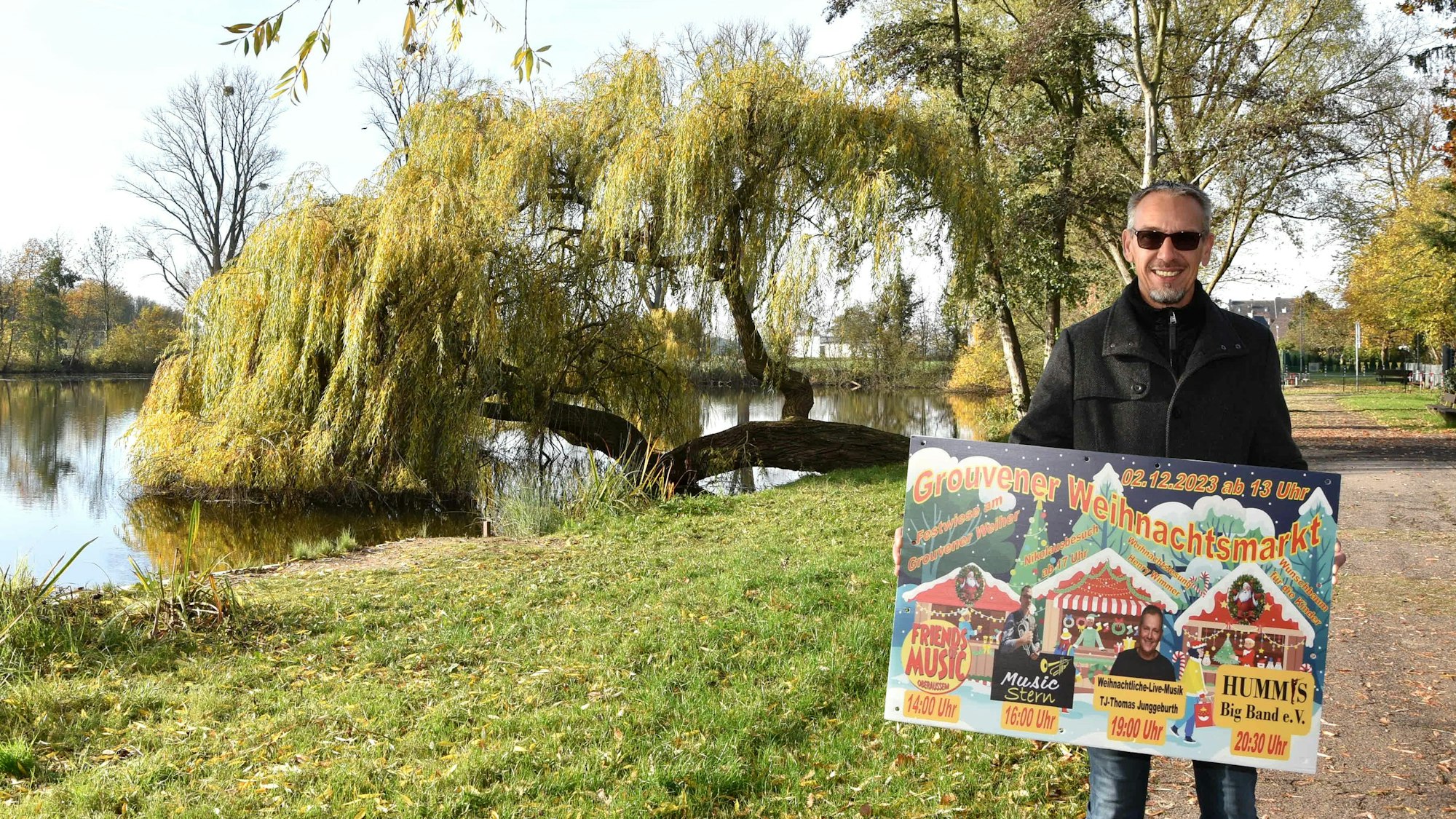 Zu sehen ist Ralf Gohrbandt, der vor dem Grouvener Weiher steht und ein Weihnachtsmarkt-Plakat in den Händen hält.