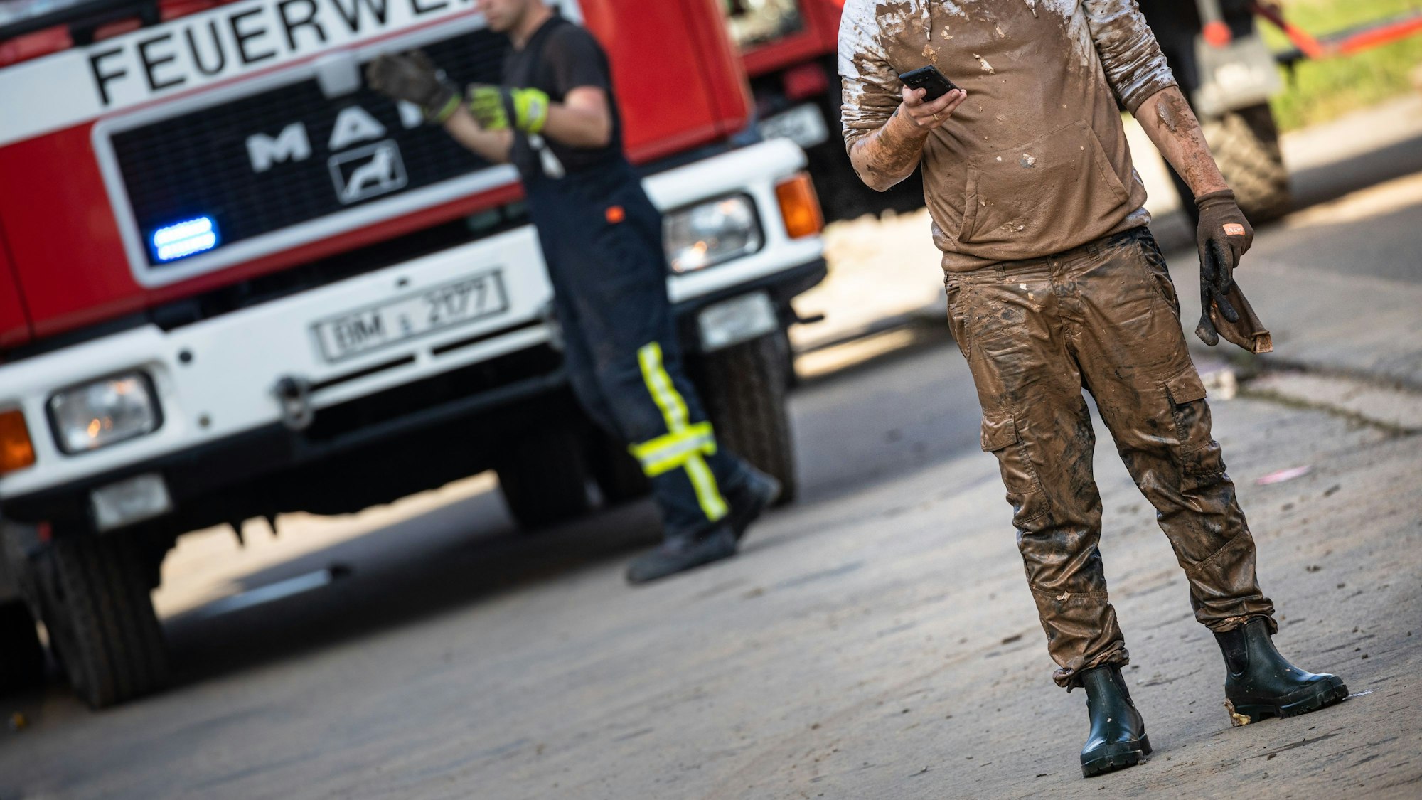 17.07.2021, Erftstadt: Ein Helfer, dessen Kleidung total verschlammt ist, blickt im Ortsteil Bliesheim auf sein Handy. Nach dem Hochwasser beginnen die Aufräumarbeiten. Starkregen hatte am Mittwoch zu schwersten Überschwemmungen und Hochwasser geführt. Der Rhein-Erft-Kreis hat Katastrophenalarm ausgelöst. Foto: Matthias Heinekamp