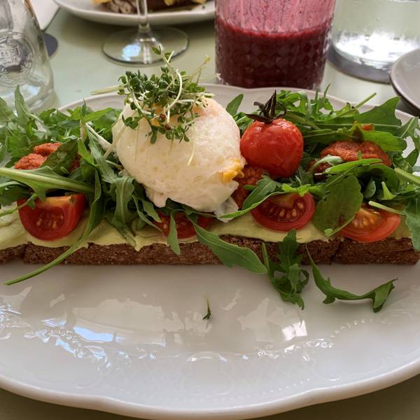 Auf einem weißen Teller liegt ein belegtes Brot mit Avocado-Creme, Tomate, Rucola, Mozzarella-Kugel und Kresse-Sprossen.