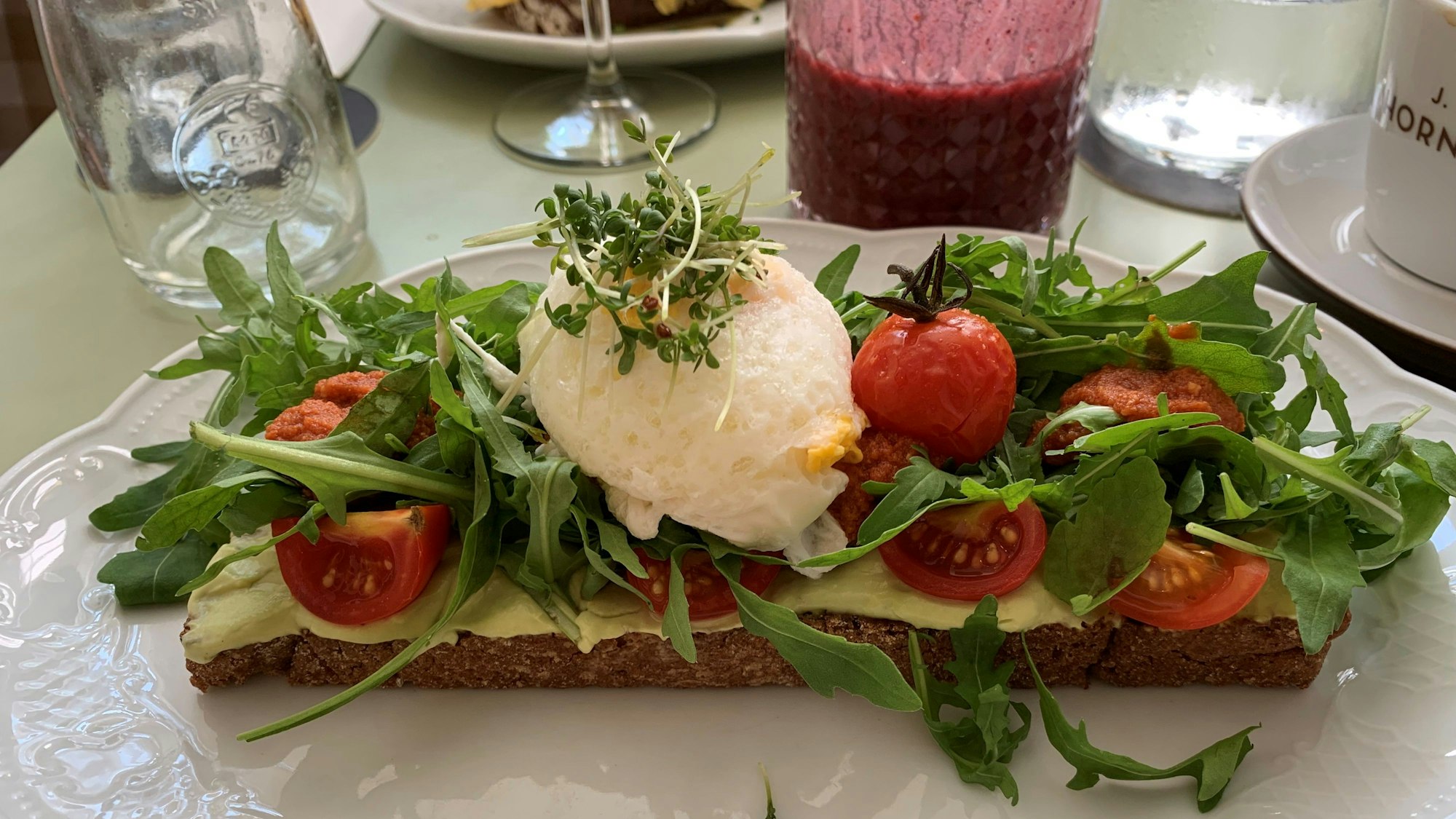 Auf einem weißen Teller liegt ein belegtes Brot mit Avocado-Creme, Tomate, Rucola, Mozzarella-Kugel und Kresse-Sprossen.