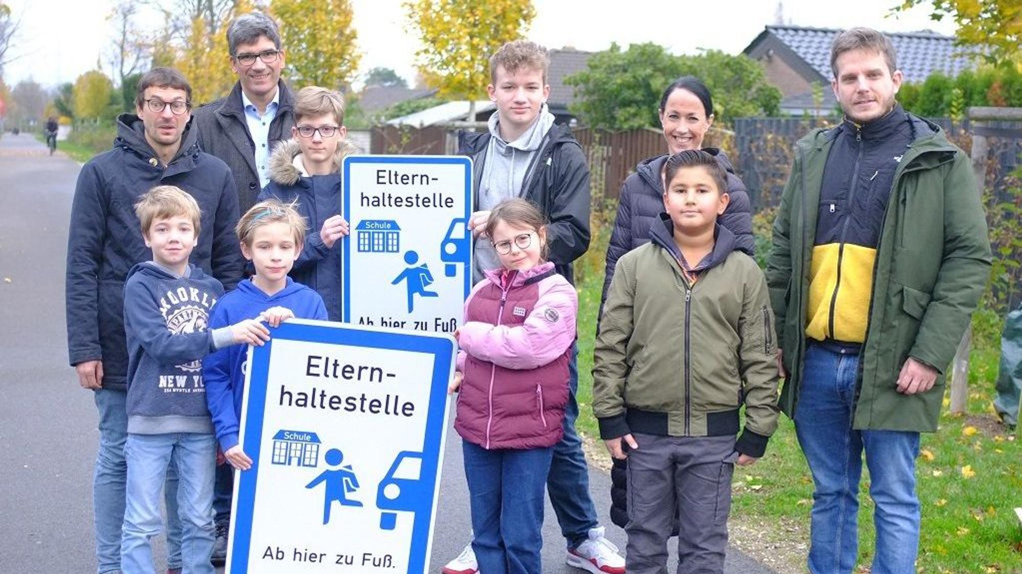 Auf dem Foto Kinder und Jugendliche, Lehrer und Erwachsene zu sehen. Die Kinder halten Schilder mit der Aufschrift „Elternhaltestelle - Ab hier zu Fuß"