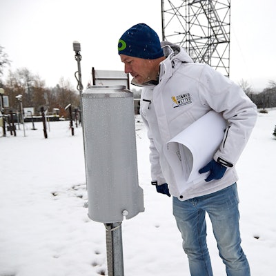 Im Vordergrund schaut Dr. Karsten Brandt in das zylindrische Niederschlagsmessgerät, im Hintergrund das Gerüst der verschneiten Wetterstation.
