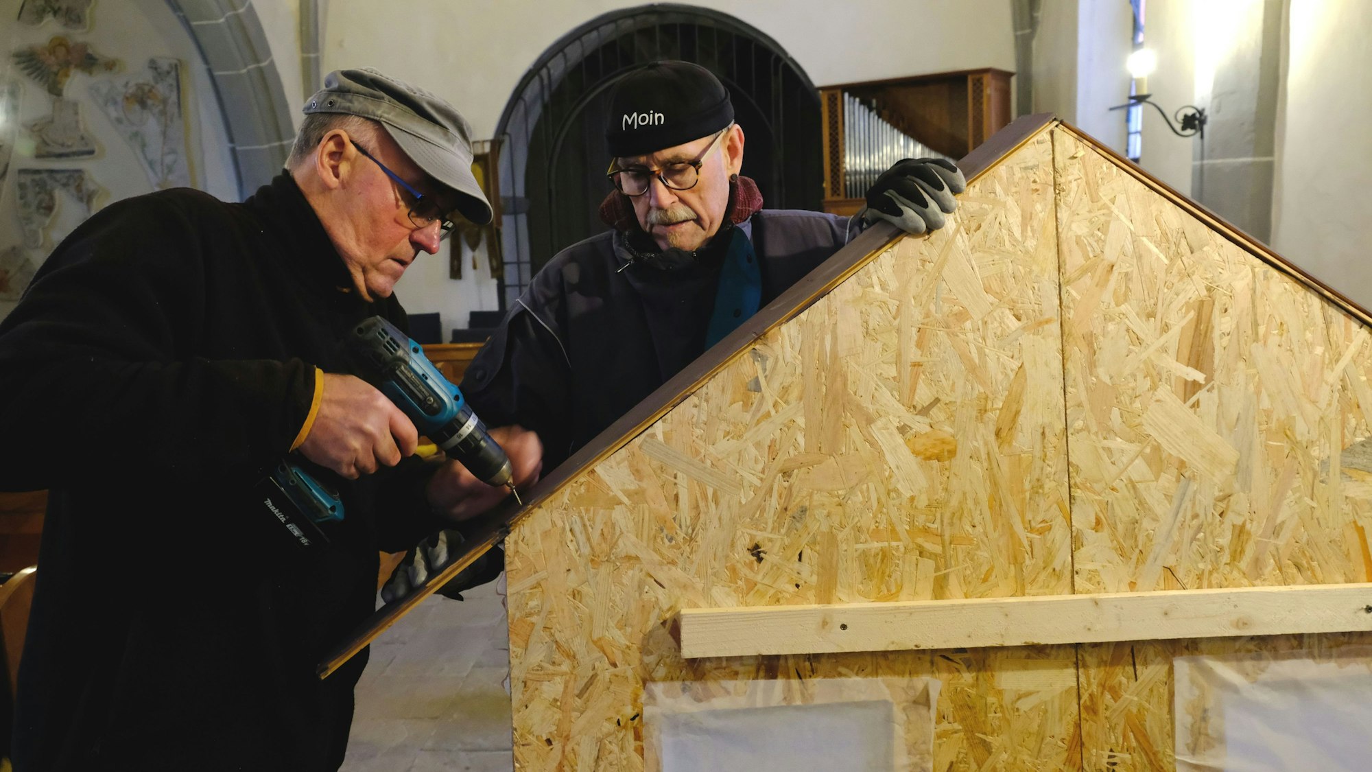 Die Krippenbauer Georg Lünenbach und Dieter Schwinning hantieren mit einem Akkuschrauber an einem aus OSB-Platten gefertigten Haus einer Weihnachtskrippe.