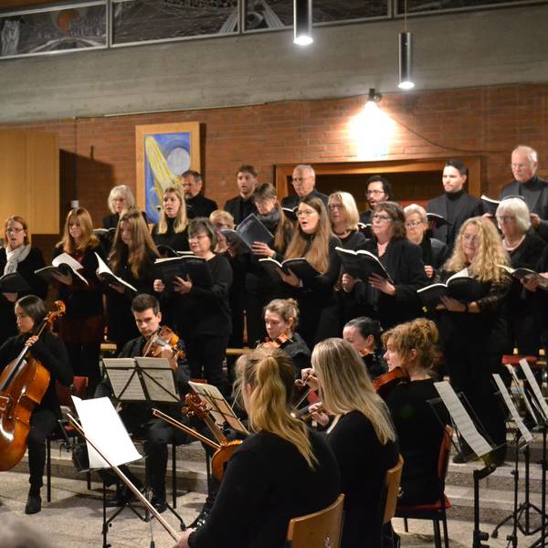 Ein Chor singt in schwarzer Kleidung, davor sitzt ein keines Orchester.