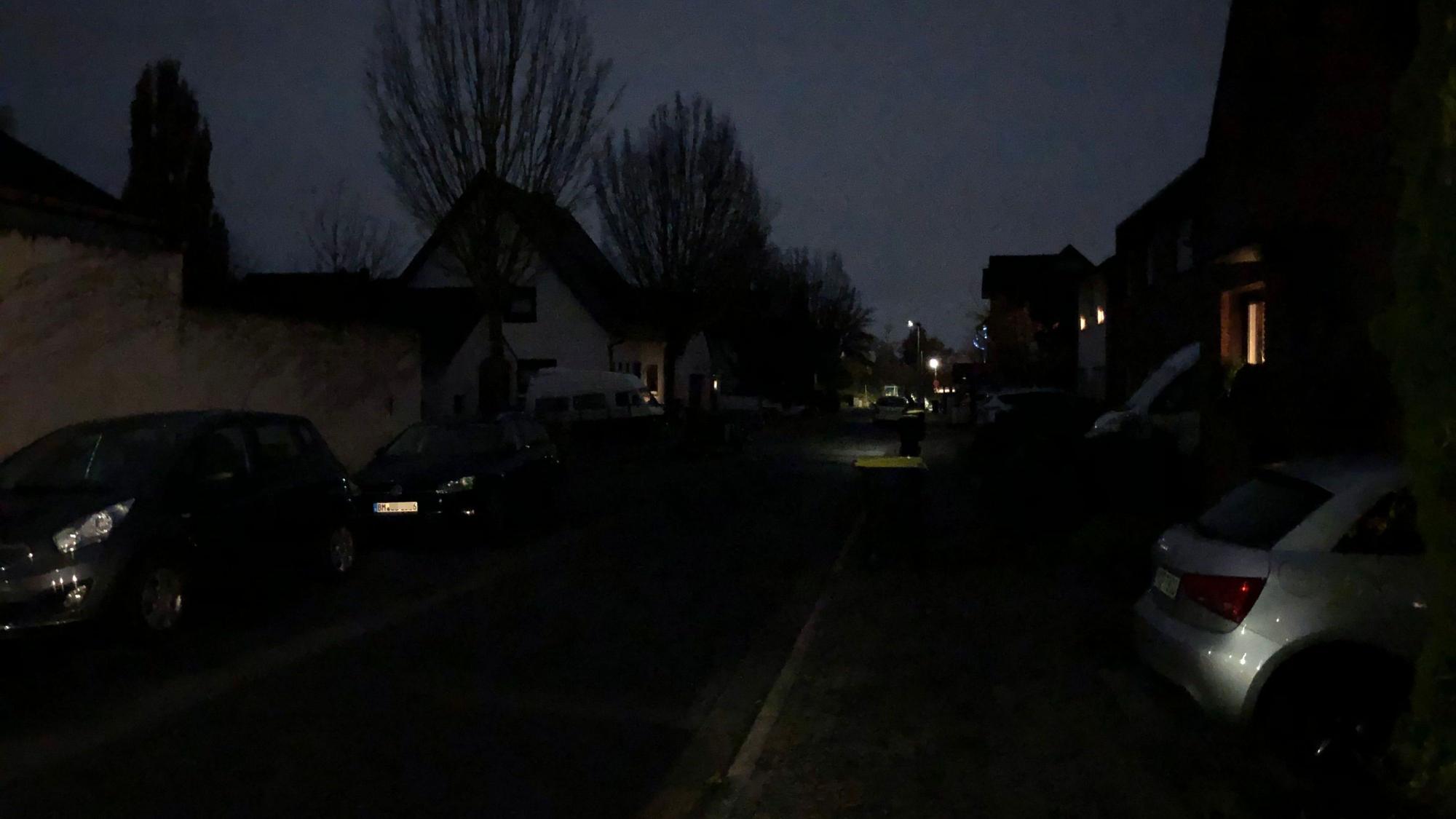 Ein Straßenzug ist nur sehr schwach beleuchtet, es sind nur Umrisse von Häusern, Bäumen und Autos zu sehen.