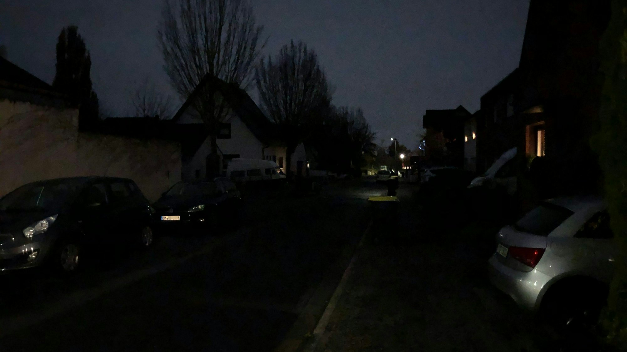 Ein Straßenzug ist nur sehr schwach beleuchtet, es sind nur Umrisse von Häusern, Bäumen und Autos zu sehen.