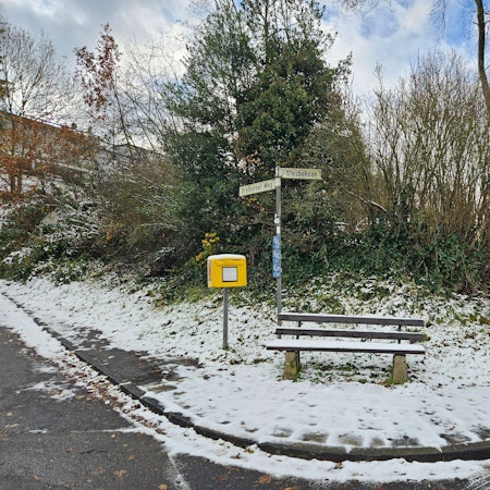 Zu sehen ist eine verschneite Bank mit Briefkasten der Post an einer Straßenkreuzung.