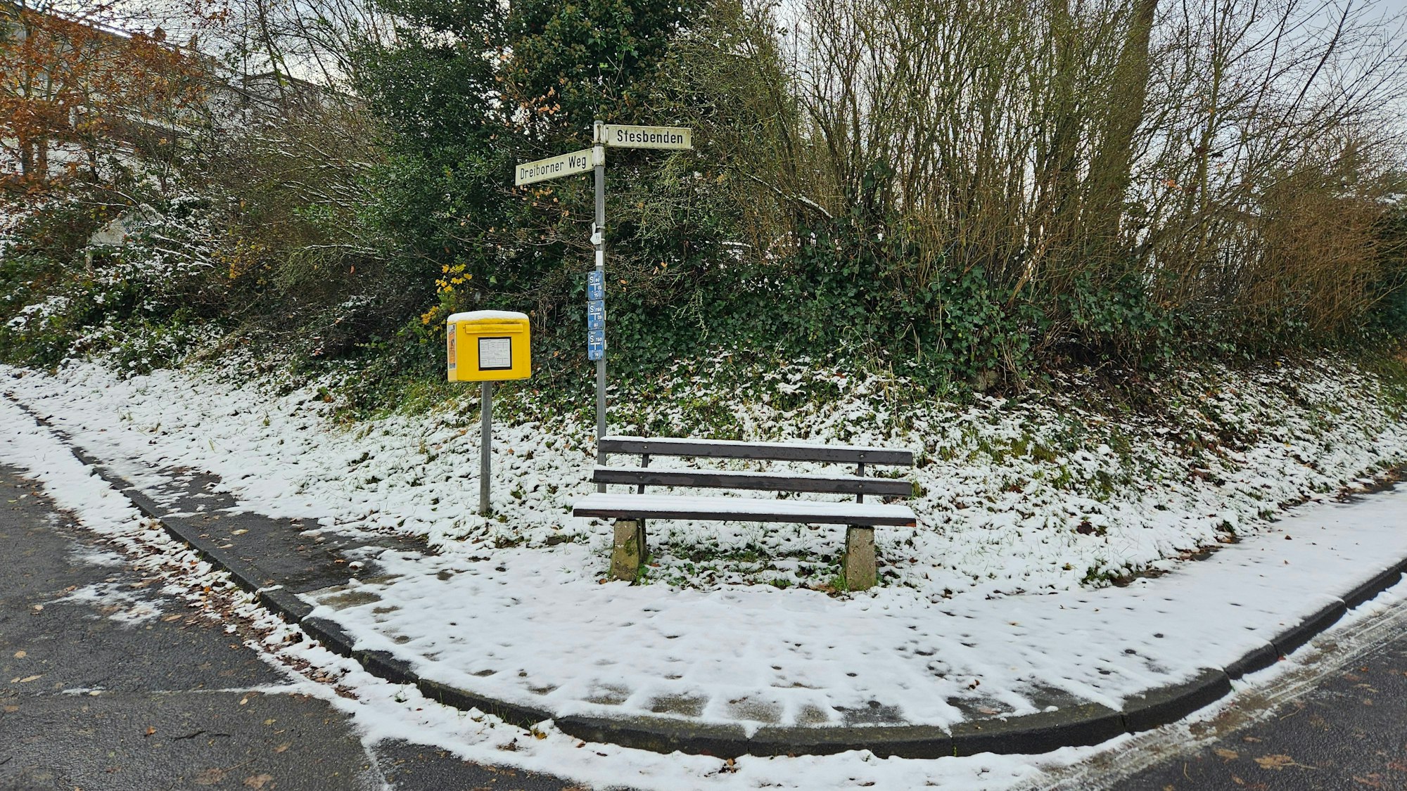 Zu sehen ist eine verschneite Bank mit Briefkasten der Post an einer Straßenkreuzung.