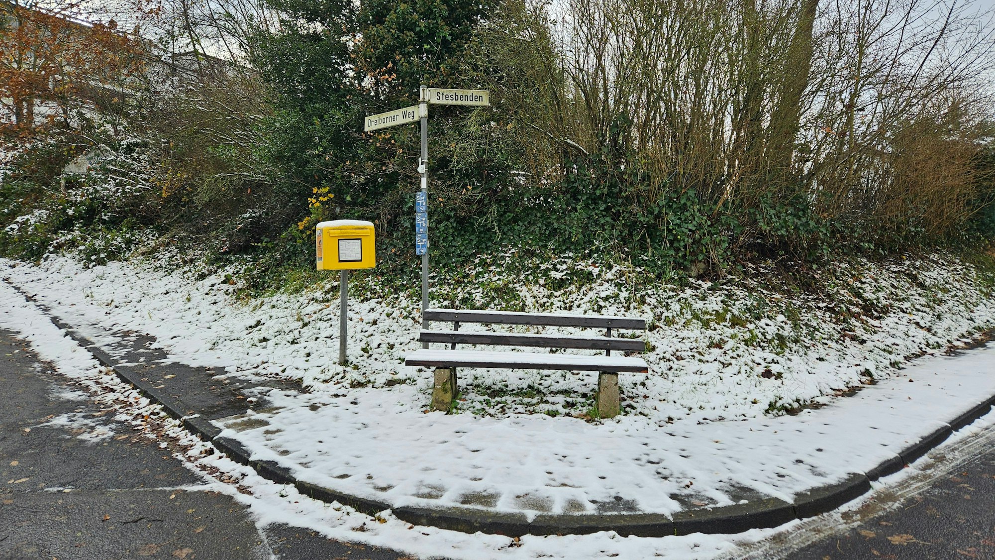 Zu sehen ist eine verschneite Bank mit Briefkasten der Post an einer Straßenkreuzung.