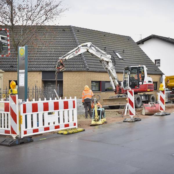 Arbeite in Orangen Jacken stehen an einem abgesperrten Stück Straße, ein Bagger und ein Lastwagen sind ebenfalls zu sehen.