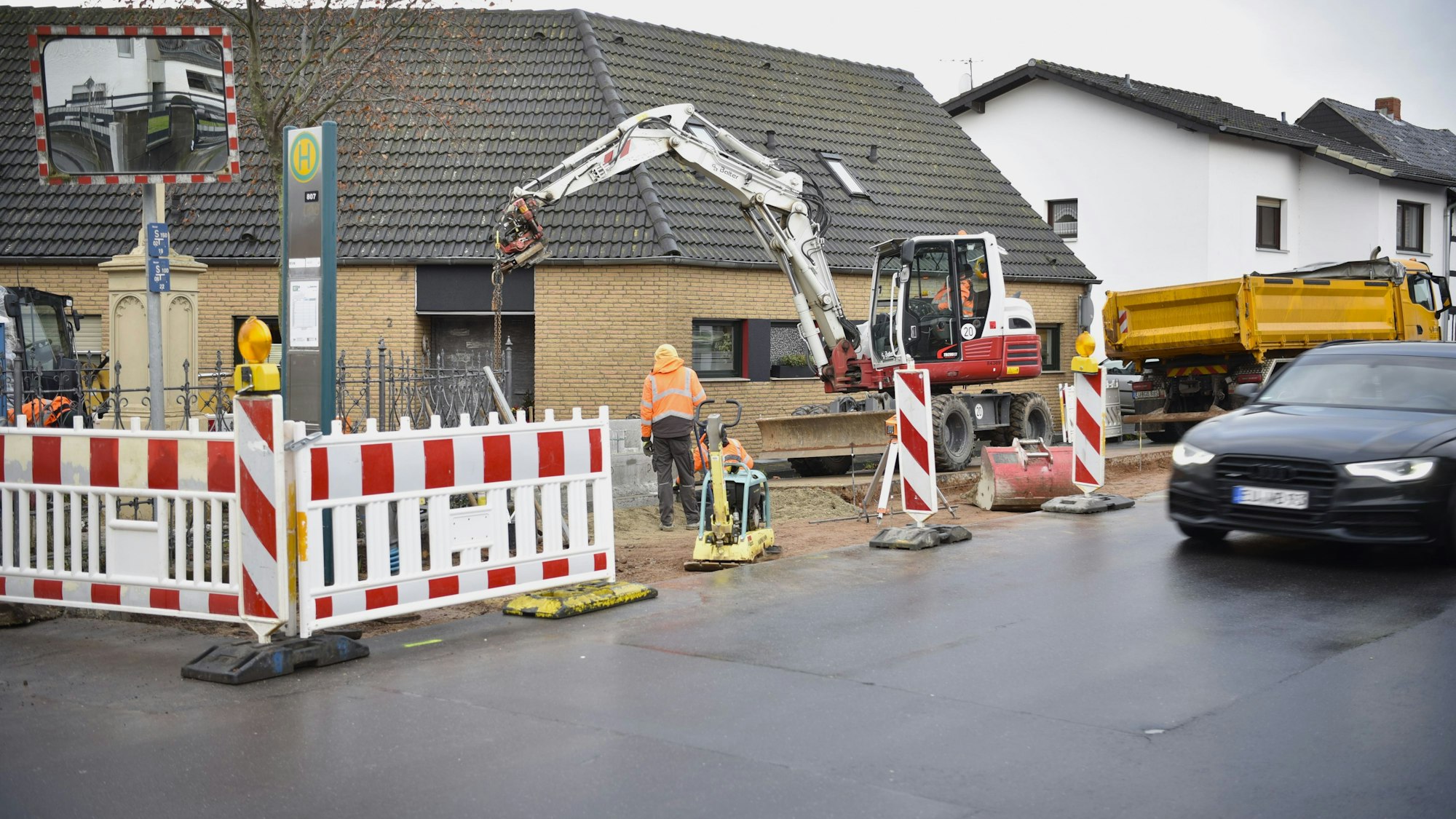 Arbeite in Orangen Jacken stehen an einem abgesperrten Stück Straße, ein Bagger und ein Lastwagen sind ebenfalls zu sehen.
