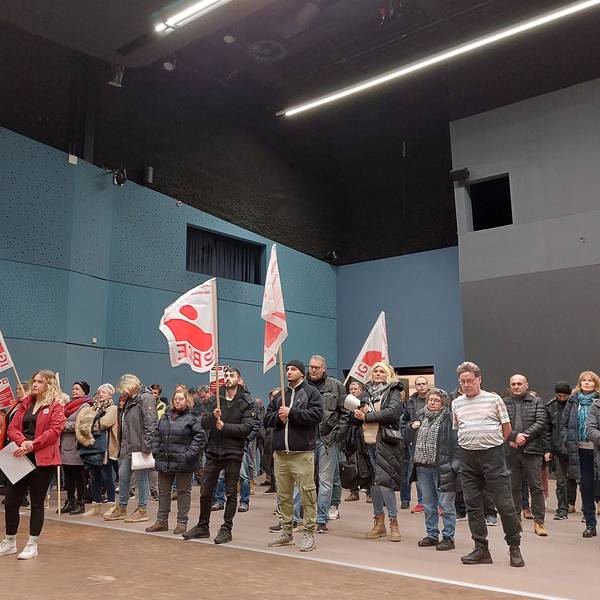 Männer und Frauen stehen in einer Veranstaltungshalle. Einige haben weiß-rote Fahnen ihrer Gewerkschaft dabei. Die Stimmung ist ernst.
