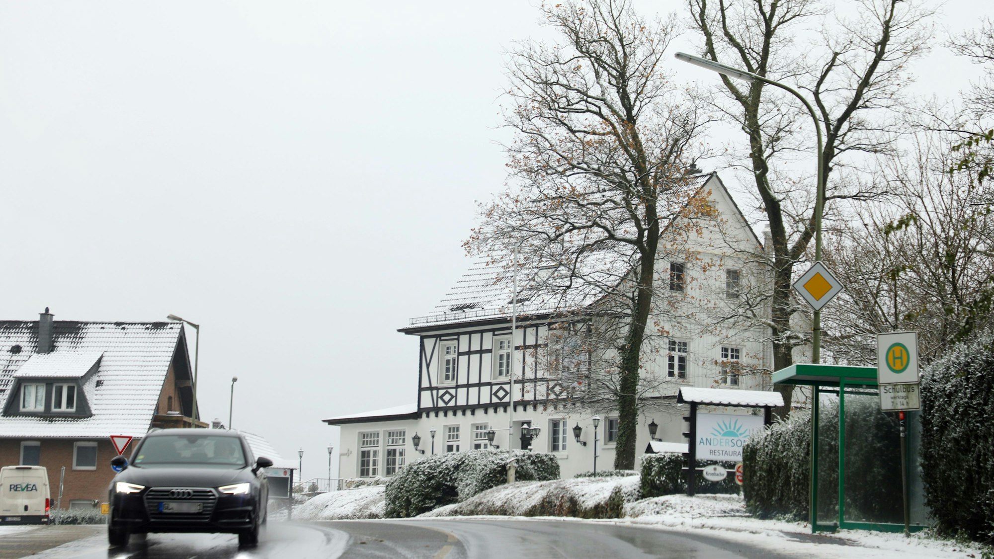 Ein Auto fährt auf einer Straße mit Schneematsch, im Hintergrund ist ein verschneites Fachwerkhaus zu sehen.