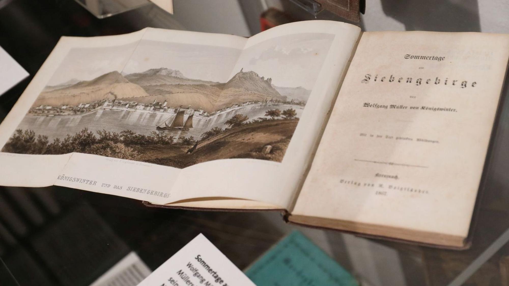 Ein aufgeschlagenes Buch mit dem Titel „Siebengebirge“ zeigt eine Ansicht der Landschaft mit Drachenfels und Rhein.