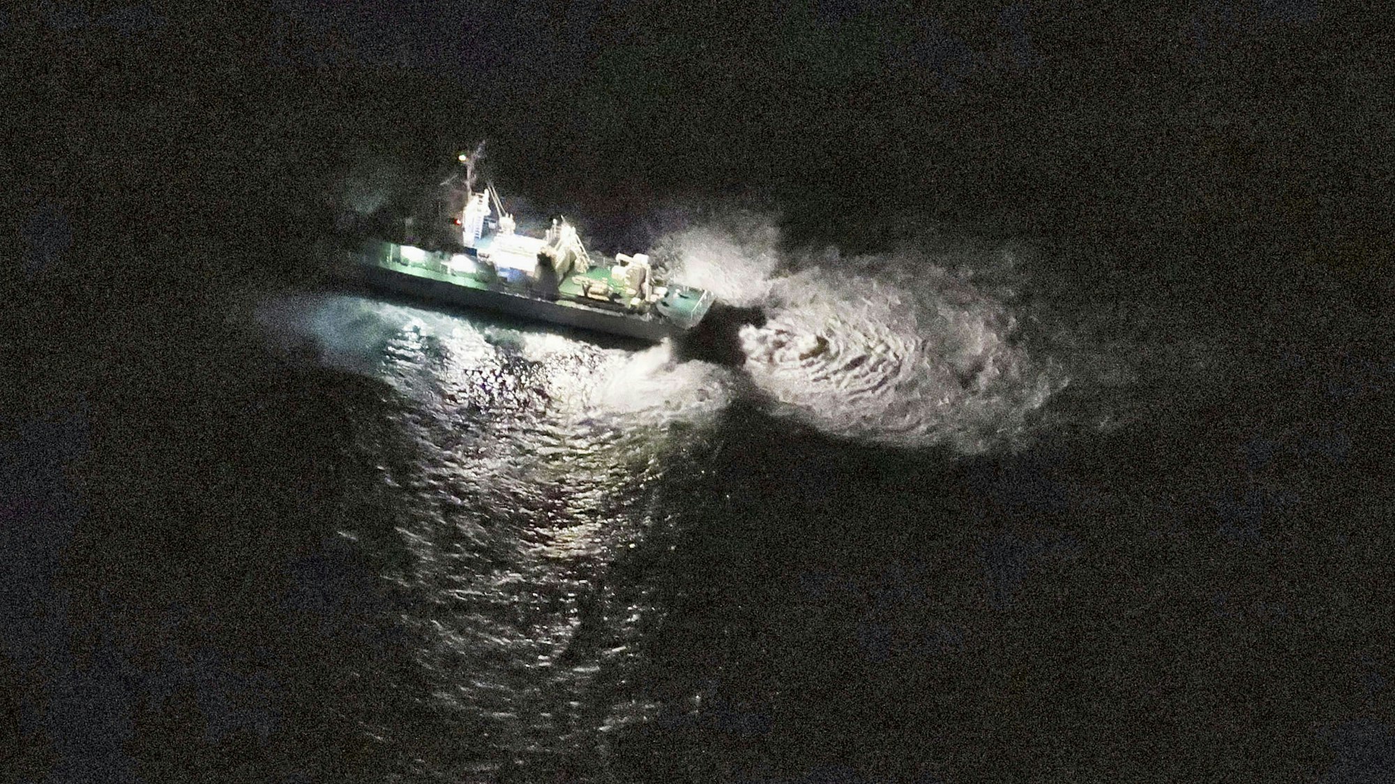 Eine Luftaufnahme zeigt ein Schiff der Küstenwache, dass auf dunkles Wasser leuchtet, in dem dadurch Wellen erkennbar werden.