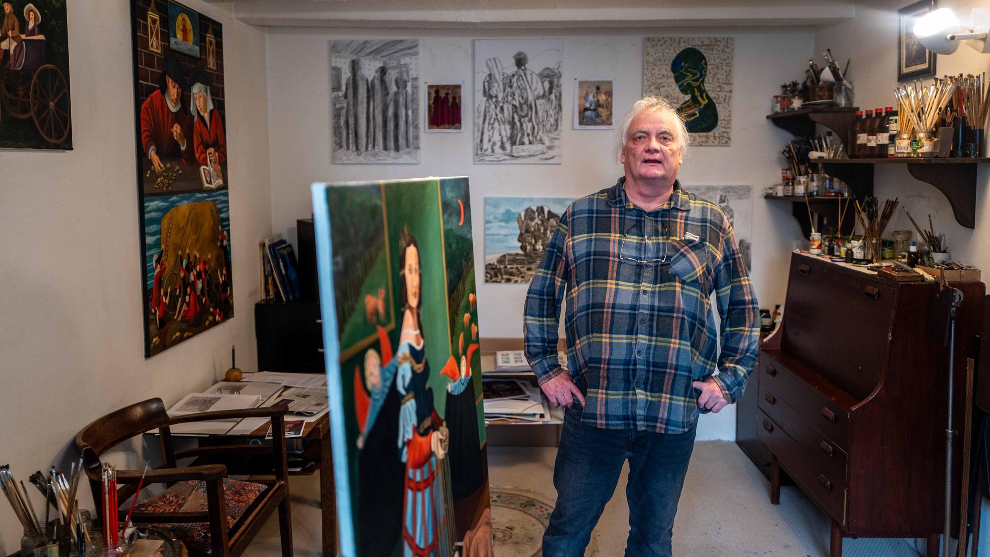 Michael Münch steht in seinem Atelier, an den Wänden hängen Bilder, in einer Ecke steht eine Kommode, auf der viele Pinsel und Malutensilien lagern.
