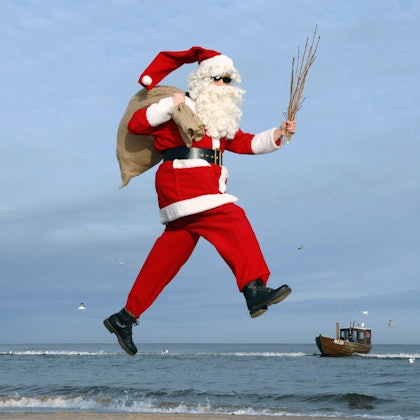 Ein Mann im Nikolauskostüm mit Sonnenbrille hüpft an einem Strand.