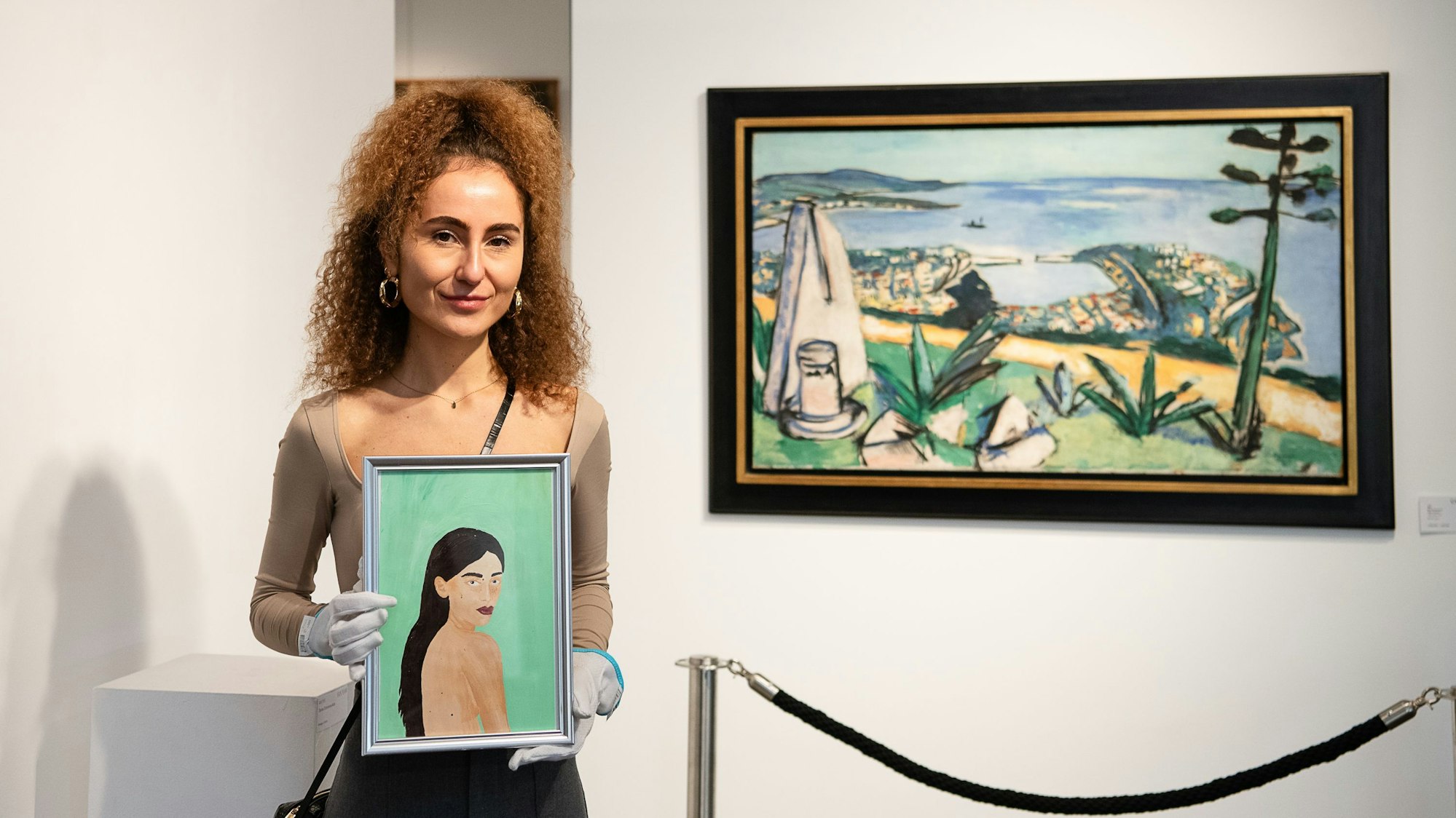 Die Künstlerin hält ihr Bild mit weißen Handschuhen in die Kamera. Neben ihr das Gemälde „Monte Carlo“ von Max Beckmann.