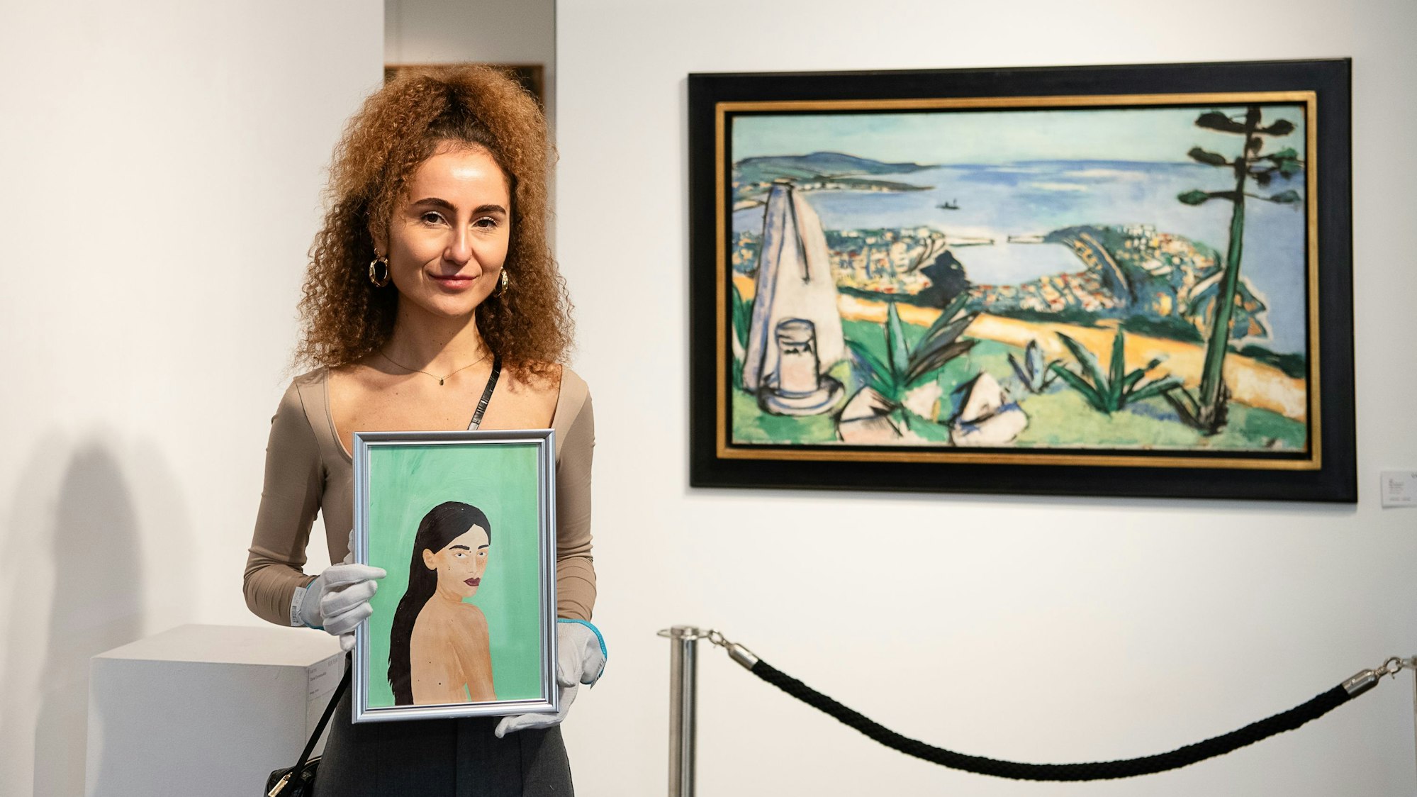 Eine junge Frau steht vor einer weißen Wand mit einem Gemälde. In ihren Händen hält sie ein kleineres Gemälde, auf dem eine andere junge Frau zu sehen ist.