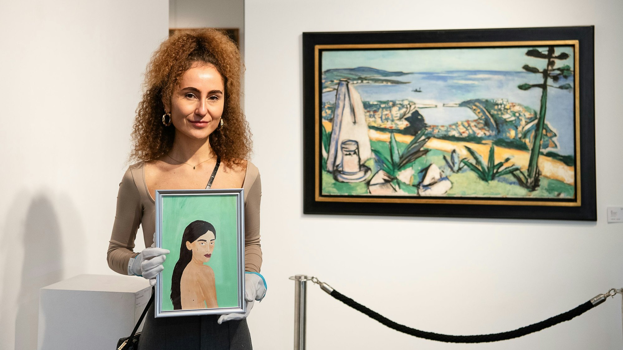Eine junge Frau steht vor einer weißen Wand mit einem Gemälde. In ihren Händen hält sie ein kleineres Gemälde, auf dem eine andere junge Frau zu sehen ist.