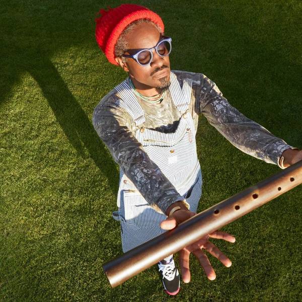 André 3000 trägt eine blau-weiß gestreifte Latzhose und eine orange Wollmütze. Er hat die Augen hinter seiner Brille geschlossen und h