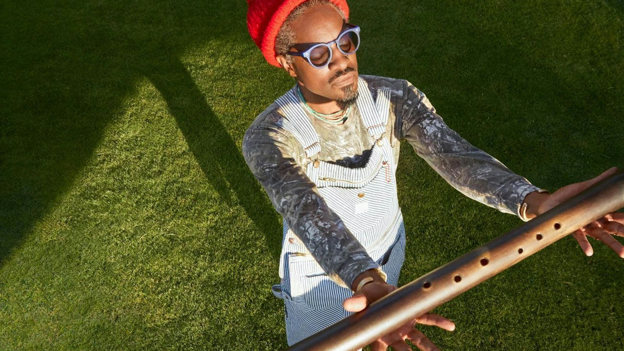 André 3000 trägt eine blau-weiß gestreifte Latzhose und eine orange Wollmütze. Er hat die Augen hinter seiner Brille geschlossen und h