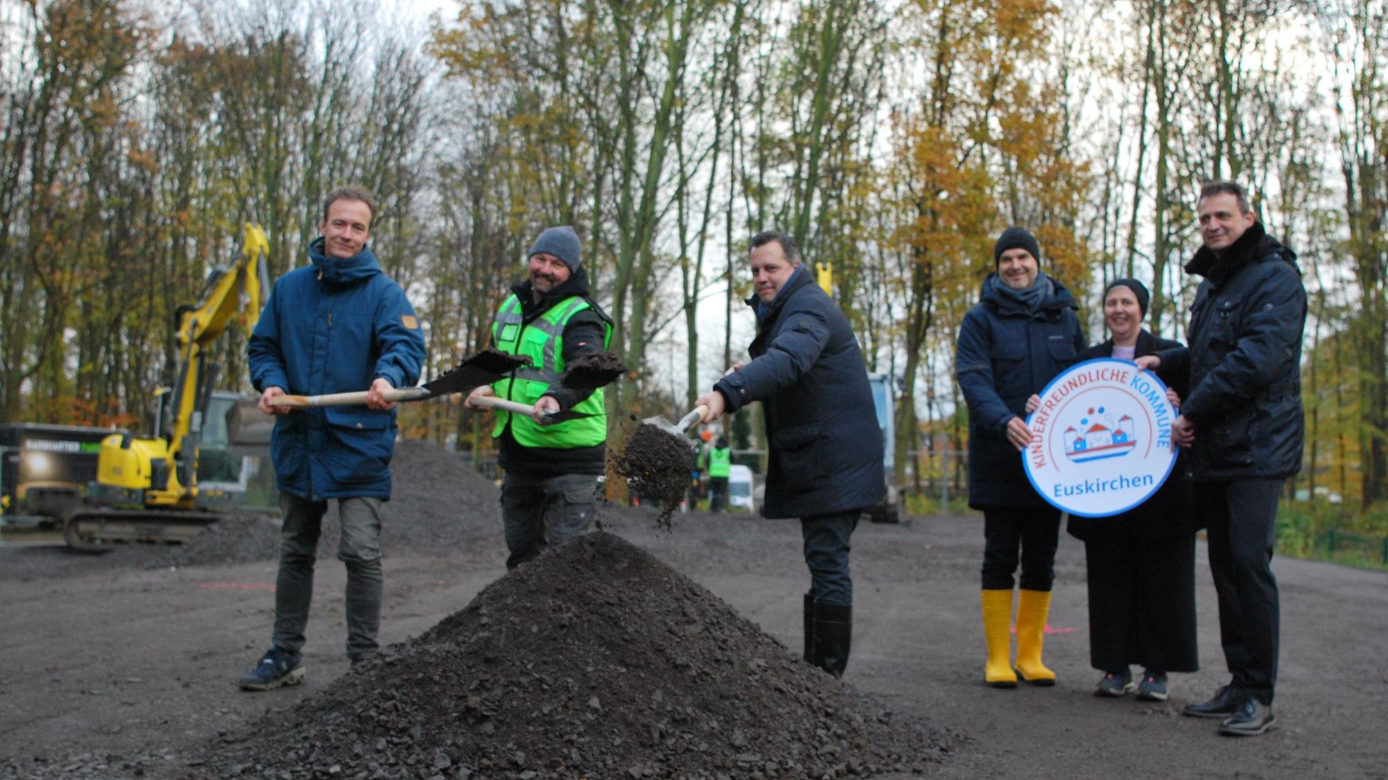 Vertreter der Stadt Euskirchen und der Firma Radquartier werfen mit Spaten Splitt in die Luft. Neben ihnen stehen drei städtische Bedienstete mit einem symbolischen Siegel, das Euskirchen als kinderfreundliche Kommune ausweist.