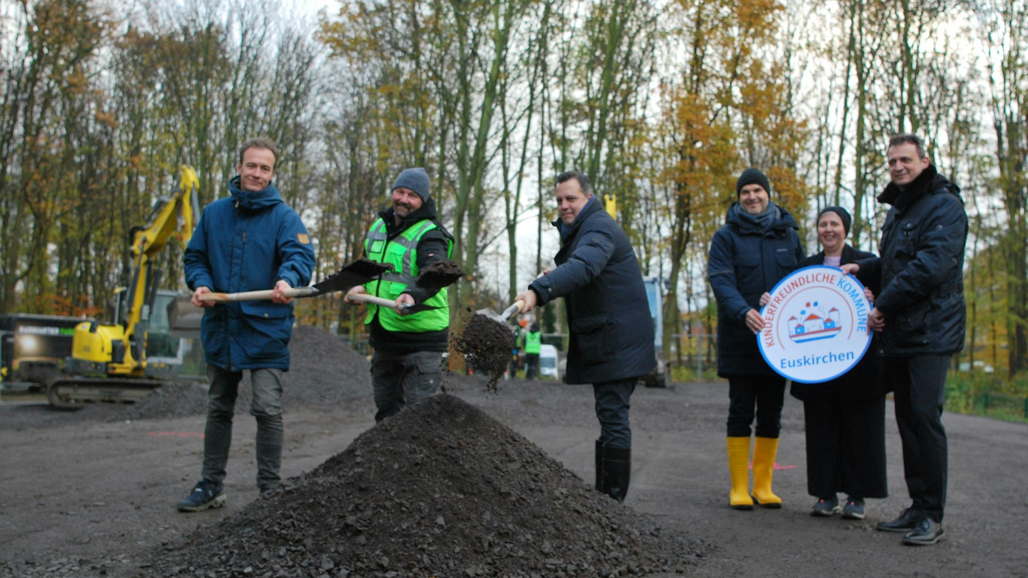 Vertreter der Stadt Euskirchen und der Firma Radquartier werfen mit Spaten Splitt in die Luft. Neben ihnen stehen drei städtische Bedienstete mit einem symbolischen Siegel, das Euskirchen als kinderfreundliche Kommune ausweist.