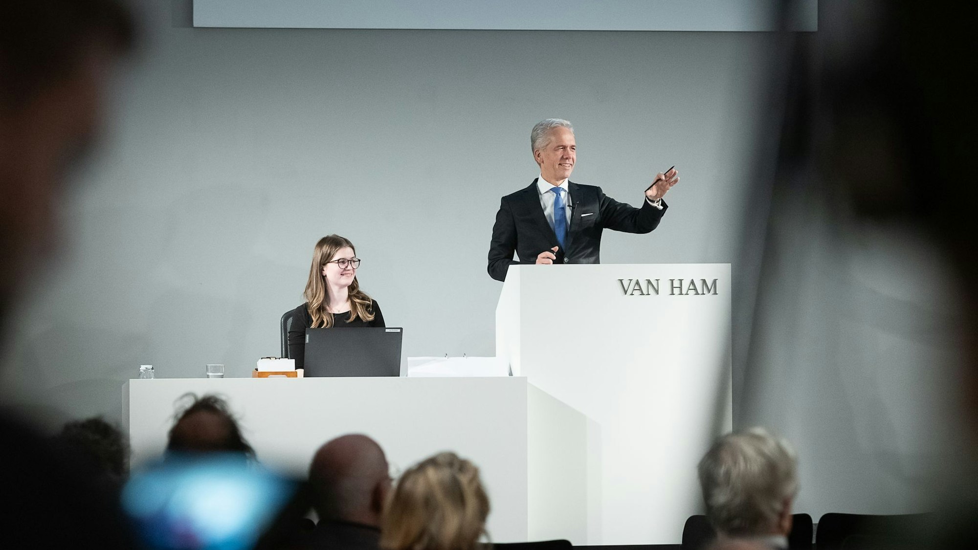 Ein Mann steht hinter einem Pult und hebt eine Hand. Neben ihm eine Frau am Computer. Davor stehen