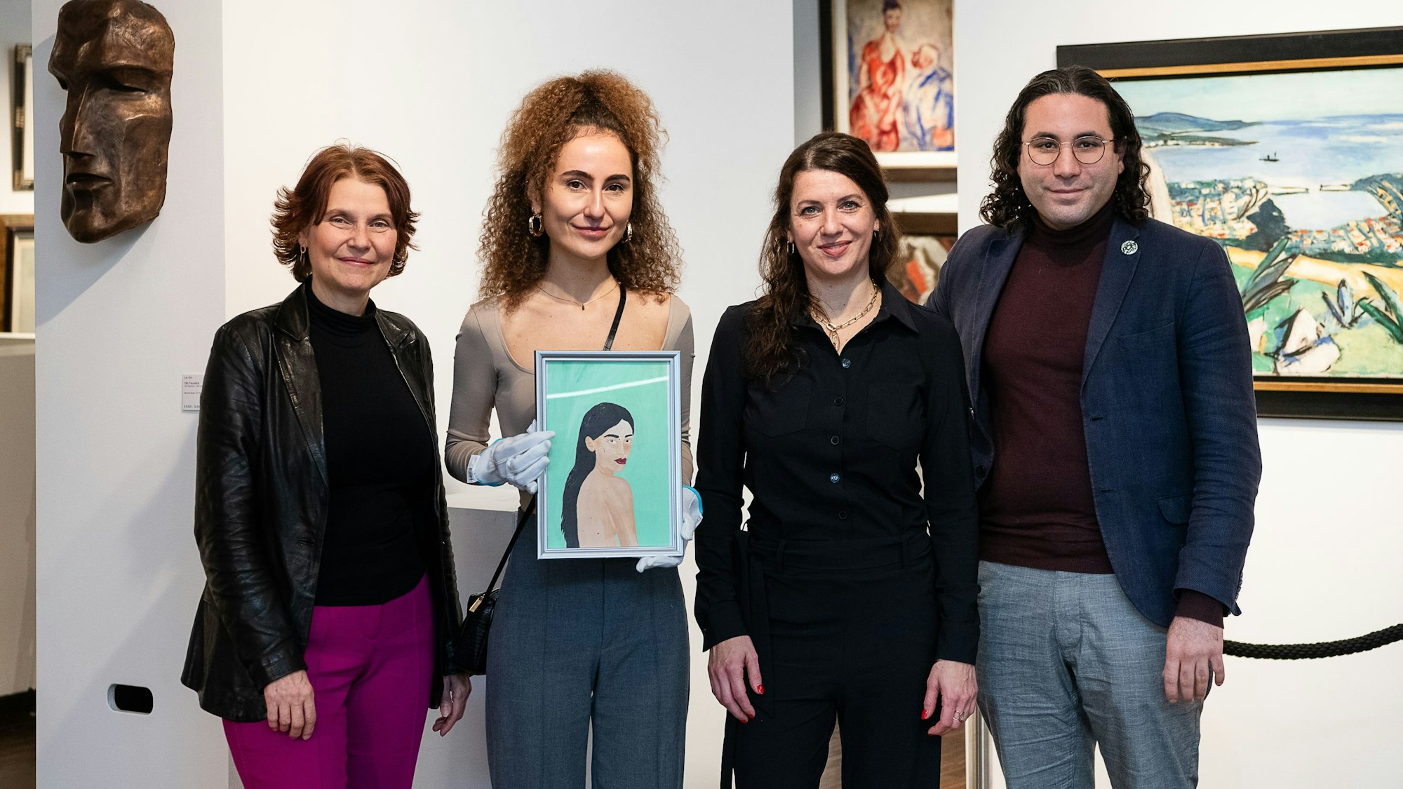 Drei Frauen verschiedenen Alters und ein Mann stehen vor einer weißen Wand mit Skulpturen und Gemälden. Die jüngste Frau hält ein Gemälde in den Händen.